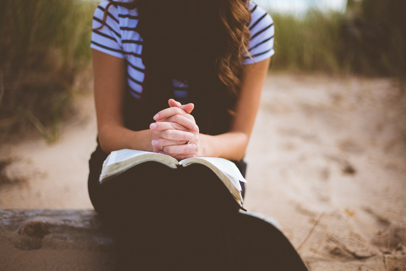 woman sitting on brown bench while reading book hands folded in prayer over a bible